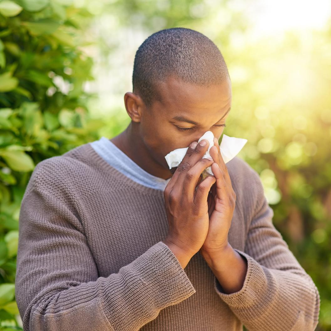 Young boy outdoors holding a tissue to his nose, appearing to have allergies or a cold, with blurred green foliage in the background.